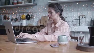 集中精力的年轻女子坐在厨房的木桌旁，在她的<strong>银色笔记本</strong>电脑上用茶或